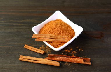 Koyu kahverengi ahşap arka planda beyaz bir kasede sopalarla yer tarçın tozu.