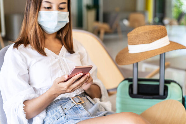 Молодая женщина в медицинских масках с багажом с помощью смартфона во время ожидания, чтобы проверить в отеле, Новая концепция нормального образа жизни путешествия