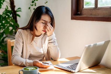 Yorgun ve stresli genç Asyalı kadın sabah bir fincan kahve ve kahvaltıyla iş yerinde oturuyor.