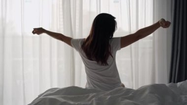 Genç Asyalı kadın yatakta oturuyor ve sabahları kendini evinde dinlendiriyor.