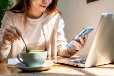 Elinde kahve fincanı olan genç bir kadın cep telefonu kullanıyor ve kahve dükkanında dizüstü bilgisayarla çalışıyor.