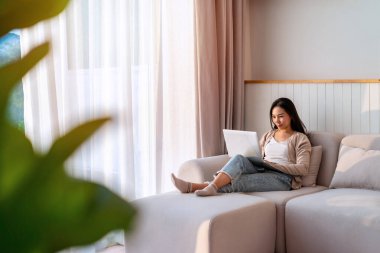Genç Asyalı kadın sabahları evdeki kanepede dizüstü bilgisayar kullanıyor.