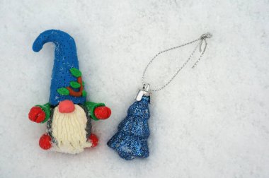 Kar zemininde plastikinden yapılmış bir cüce figürü. Yanında küçük bir oyuncak Noel ağacı var..