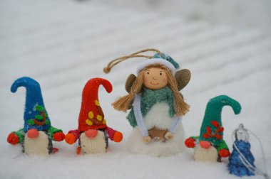 Beyaz karlı arka planda üç cüce figürü. Oyuncak bir melek ve Noel ağacının yanında.