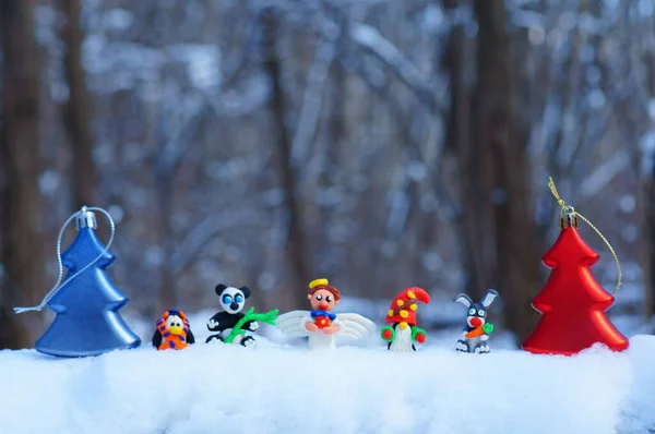 Figurines of fairy-tale characters in the winter forest. An angel with a gift, a dwarf, a rabbit with a carrot, a panda, a penguin in a scarf. There are toy Christmas trees nearby.