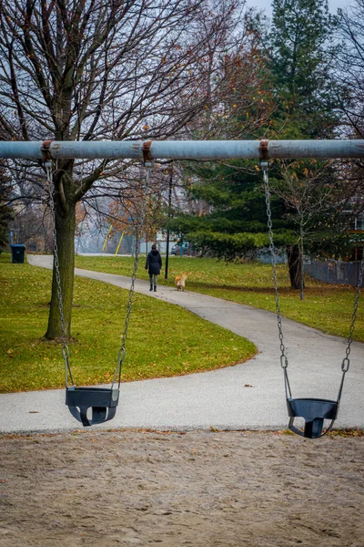 Kışlık ceketli bir kadın köpeğini boş bir çocuk parkında gezdirir. — Stok fotoğraf