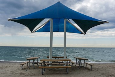 Pickering, Ontario, Kanada - Arka planda dalgalı göl suyu ve bulutlu gökyüzü olan mavi bir gölün altında sahil piknik masaları