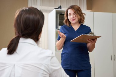 Doktor genç bayan hastayla konuşuyor ve yanında durup notlar alıyor.