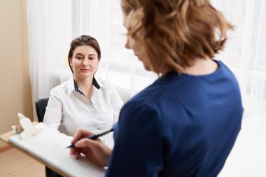 Doktor genç bayan hastayla konuşuyor ve yanında durup notlar alıyor.