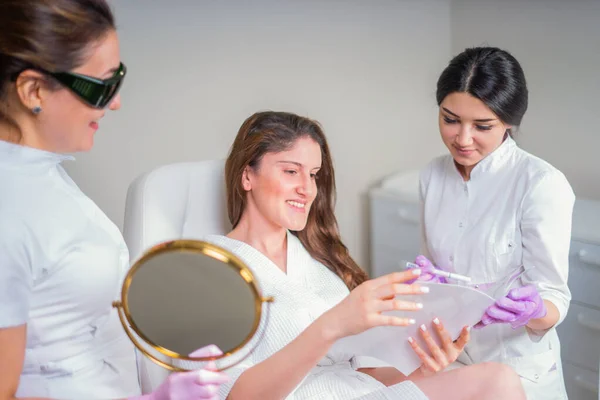 Twee cosmetologen raadplegen vrouwelijke cliënt die kijkt naar spiegel in schoonheidskliniek — Stockfoto