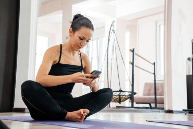 Telefonla antremandan sonra dinlenen kadın. Yoga yaptıktan sonra spor yapan genç bir kadın, egzersiz yapmaya ara vermek yoga minderinde dinlenmek, cep telefonuyla mesajlaşmak, akıllı telefon tutmak.