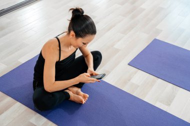 Telefonla antremandan sonra dinlenen kadın. Yoga yaptıktan sonra spor yapan genç bir kadın, egzersiz yapmaya ara vermek yoga minderinde dinlenmek, cep telefonuyla mesajlaşmak, akıllı telefon tutmak.