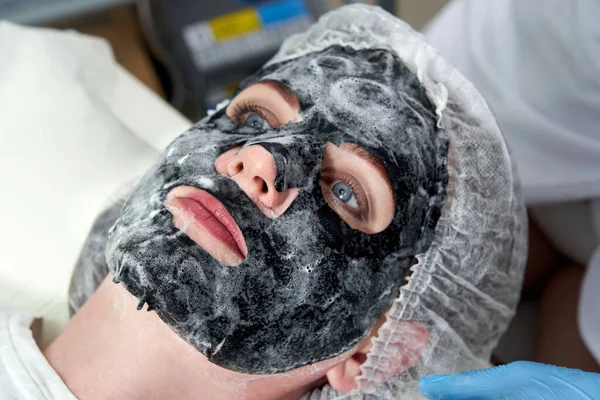Wanita muda dengan masker gelembung oksigen hitam di wajahnya di salon kecantikan — Stok Foto