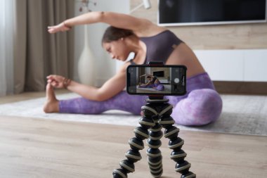 Genç bayan blogcu oturma odasında yoga yaparken spor videosu kaydediyor.