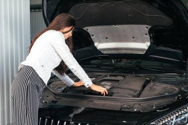 Garajında kaputu açık duran genç bir kadın arabanın kaputunun altına bakıyor.