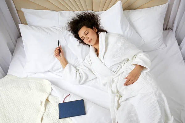 Gündüz vakti yatakta uyuyan genç bir kadının tepeden bakışı. Yanında defter ve elinde kalem var. — Stok fotoğraf