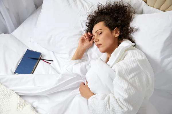 Yatakta uyuyan kıvırcık kadın yanında bornoz giyerek not defteriyle uyuyor. — Stok fotoğraf
