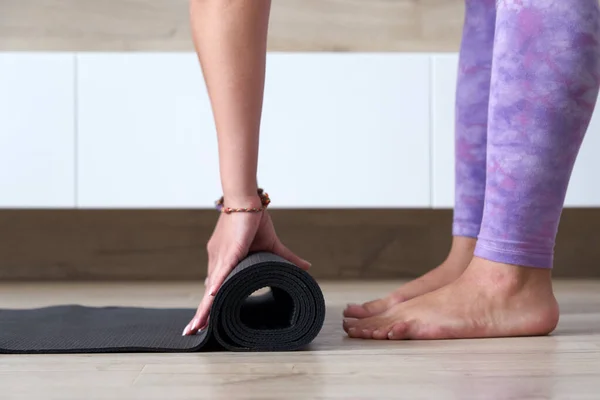 Young woman folding black yoga or fitness mat after working out at home in living room. Healthy life, yoga concepts — стоковое фото