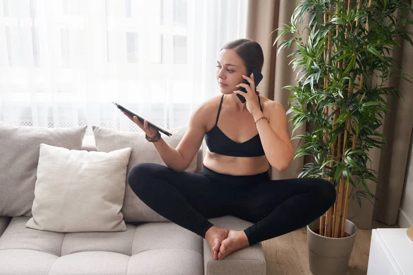 Junge Frau sitzt im Schneidersitz auf Couch in Lotus-Pose mit Tablet und Handy zu Hause im Wohnzimmer — Stockfoto