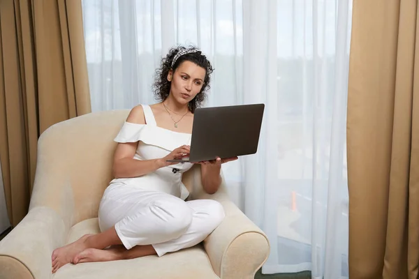 Vacker ung kvinna som använder laptop sittandes i fåtölj. Begreppet avslappnad affärskvinna i hotellrum — Stockfoto
