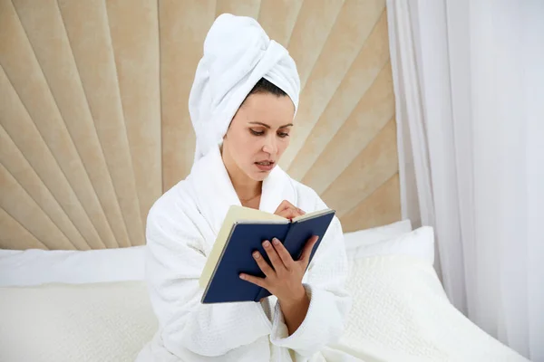 Young woman sits on bed wearing bathrobe writing notes in diary. Blogger creats publication for blog in notebook