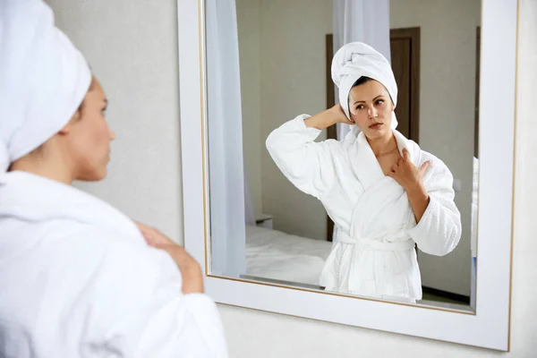 Mujer joven con albornoz y toalla en la cabeza mirando al espejo, por la mañana — Foto de Stock