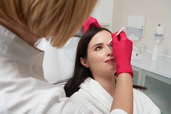 Female cosmetologist drawing line on female face with a white marker to prepare for the procedure. Facial care concept