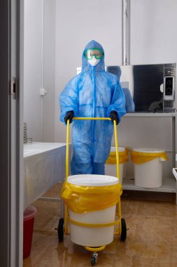 PPE koruyucu giysi, maske ve eldiven giyen bir teknoloji uzmanı araştırma laboratuarında büyük plastik kovayla çalışıyor.