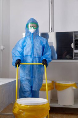 PPE koruyucu giysi, maske ve eldiven giyen bir teknoloji uzmanı araştırma laboratuarında büyük plastik kovayla çalışıyor.