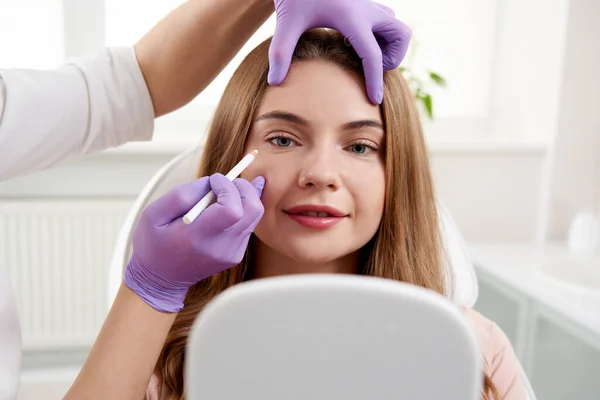 美容师在温泉美容院治疗前对女性患者面部使用面部标记。病人正在美容院照镜子 — 图库照片