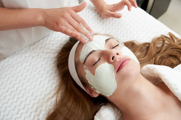 Cosmetologo che applica la maschera facciale ringiovanente su una faccia femminile in salone di bellezza. Trattamento viso concetto — Foto Stock