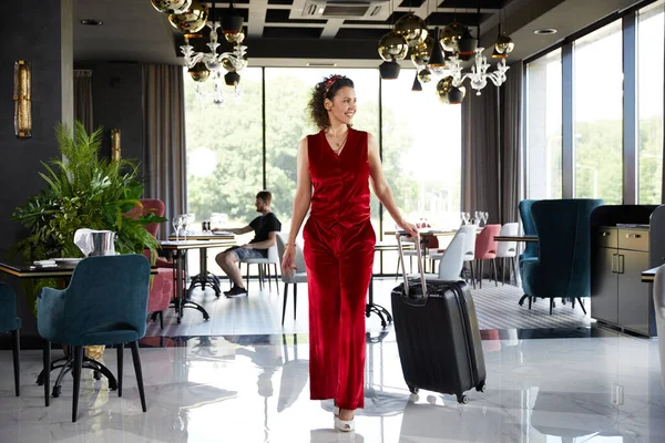 Okouzlující mladá žena v červeném sametovém obleku, která se usmívá s kufrem v hotelové hale. Cestovní koncept — Stock fotografie