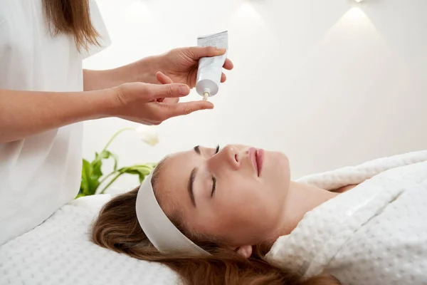 Cosmetoloog aanbrengen crème op vrouwelijk gezicht. vrouw met een gezichtsbehandeling in spa salon — Stockfoto