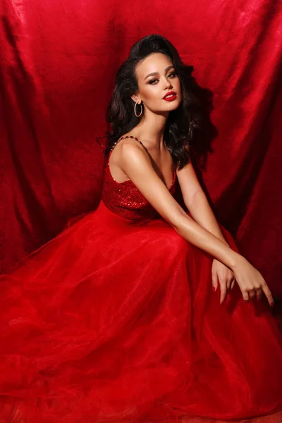 ファッション休日写真の美しいですセクシー女性とともに黒髪で豪華な赤いドレスポーズで赤いスタジオ — ストック写真