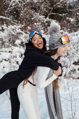 Koyu renk saçlı, sıcak spor elbiseli, kar ormanında poz veren iki güzel kadının moda fotoğrafı.