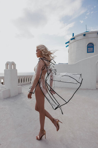 fashion outdoor photo of beautiful woman with blond hair in elegant bikini and transparent jacket posing on the sunset