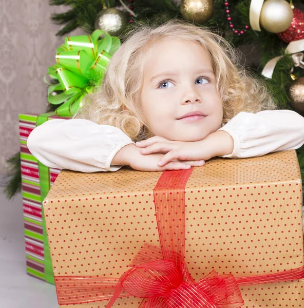 Niedliches kleines Mädchen hält eine Geschenkschachtel und sitzt neben einem Weihnachtsbaum — Stockfoto