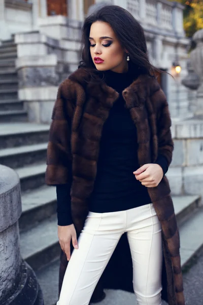 Beautiful brunette woman in luxurious fur coat posing on stairs — Stock Photo, Image