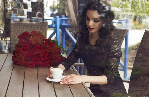 Morena sentada en caffe detrás de la ventana con gran ramo de rosas rojas — Foto de Stock