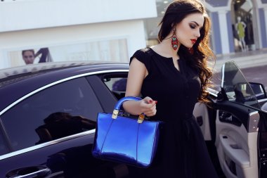 woman in elegant dress with bag, posing beside a luxurious auto