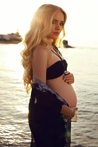 Schöne schwangere Frau mit langen blonden Haaren posiert am Strand bei Sonnenuntergang — Stockfoto