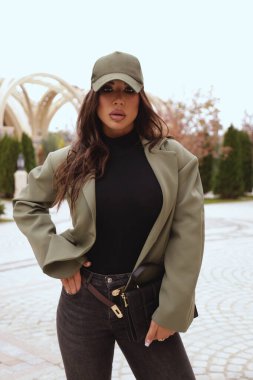 Açık hava fotoğrafında siyah saçlı, sıradan giyimli ve aksesuarlı güzel bir kadın sokakta poz veriyor.