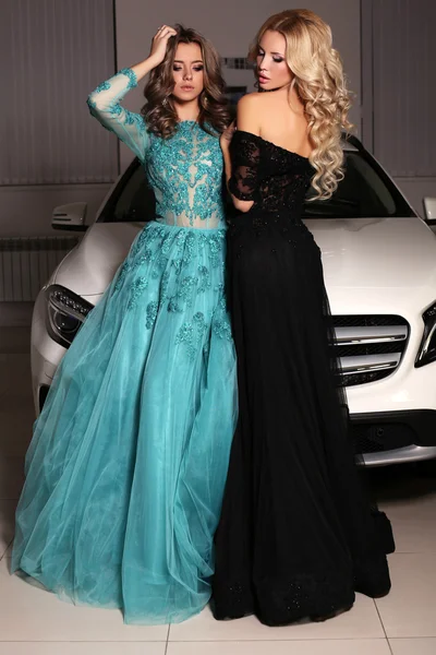 Gorgeous women with long hair wear luxurious dresses,posing beside white car — Stock Photo, Image