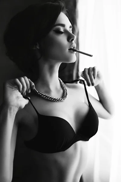 Beautiful sexy girl with dark hair wears lingerie,smoking beside a window — Stock Photo, Image