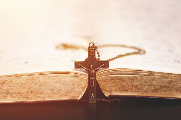  A crucifix necklace placed vertically in the middle of an open Bible, symbolizing Christian faith, sacrifice, worship, and devotion, with a warm and peaceful lighting atmosphere.