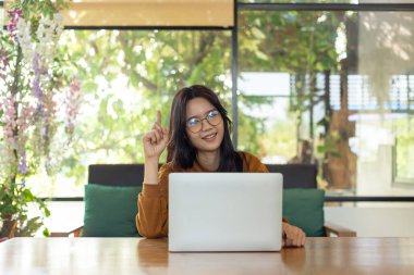 Parlak, rahat bir odada dizüstü bilgisayarla masada oturan kendine güvenen bir Asyalı kadın. Gülümsüyor ve tek parmakla yukarıya doğru işaret ediyor, bir fikri ya da sezgiyi simgeliyor. Bu resim yaratıcılık, uzak çalışma, çevrimiçi öğrenme, inovasyon gibi kavramları iletir