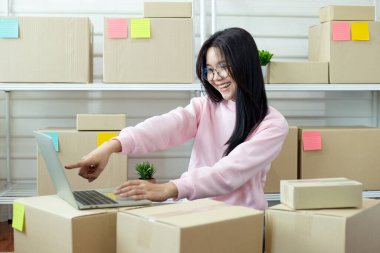 Etrafı karton kutularla çevriliyken dizüstü bilgisayarda çalışan mutlu genç Asyalı kadın. Gülümsüyor ve başarılı bir e-ticaret veya çevrimiçi perakende mağazasını temsil ederek laptop ekranına işaret ediyor. İçerik kullanımı için ideal 