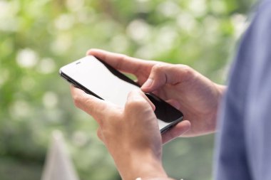 Bulanık yeşil arka planı olan, cep telefonu, teknoloji ve dijital yaşam tarzını sembolize eden akıllı bir telefonu tutan ve kullanan kişinin yakın görüntüsü.