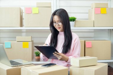 Online mağaza envanterini kutular, pano ve dizüstü bilgisayarla yöneten genç bir kadın e-ticaret, küçük işletmeler, planlama ve üretkenliği ifade ediyor..