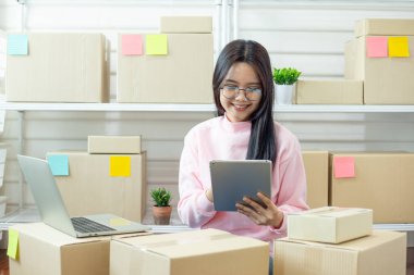 Kutular ve laptop ile çevrili online mağaza görevlerini yönetmek için tablet kullanan gülümseyen kadın, e-ticaret iş akışı, planlama ve küçük işletme verimliliğini ifade ediyor.